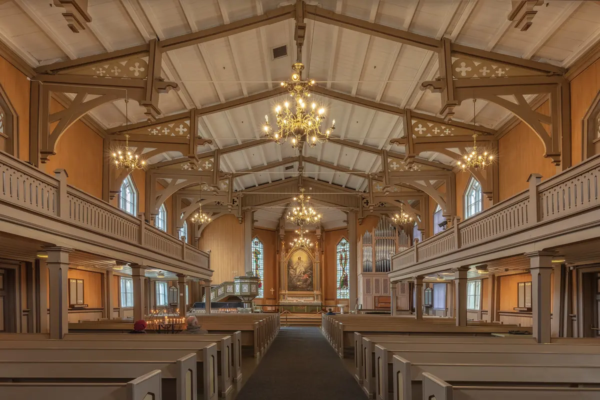 The nave of Tromsø Cathedral looking towards the altar, with pale ceiling beams, chandeliers, side galleries and rows of pews