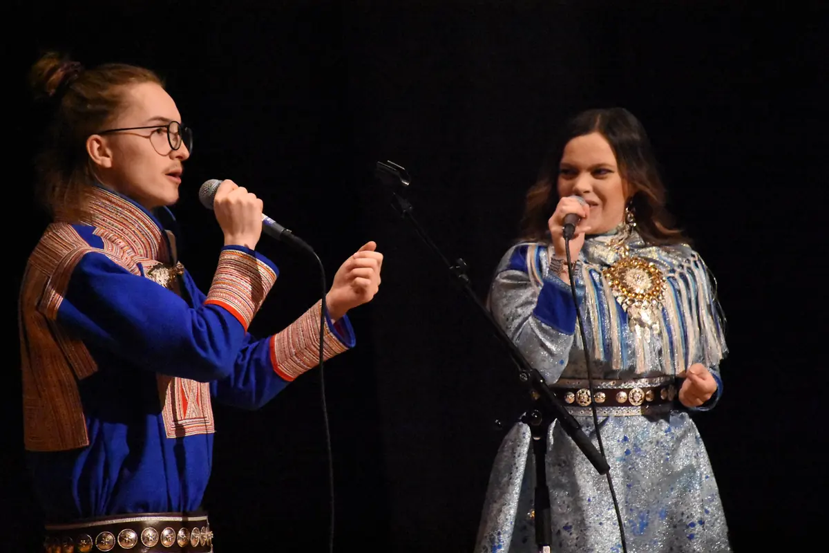 Two Sámi women in blue and silver traditional dress singing into microphones on a dark stage